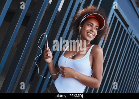 Giovane donna che indossa il tennis visor e gli auricolari in stile libero sulla strada in piedi vicino al recinto smartphone holding ascoltando musica guardando oltre sorridente h Foto Stock