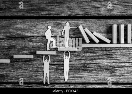 Immagine in scala di grigi di carta di piccole dimensioni persone azienda fino la caduta dei dominos stando in piedi sui gradini in legno sostenuto da due altre figure, concettuale di un business Foto Stock