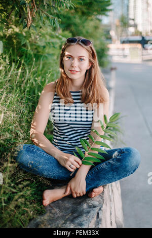 Bella bianco sorridente ragazza caucasica donna con capelli lunghi biondi e gli occhi blu che indossa striped T- shirt e jeans fuori in estate park tra verdi Foto Stock