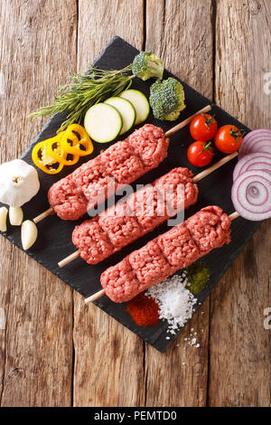 Raw, kofta kebab kofte da carni macinate su spiedini con ingredienti e verdure vicino sul tavolo. Verticale in alto vista da sopra Foto Stock