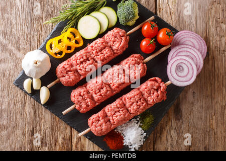 Raw, kofta kebab kofte da carni macinate su spiedini con ingredienti e verdure vicino sul tavolo. parte superiore orizzontale vista da sopra Foto Stock