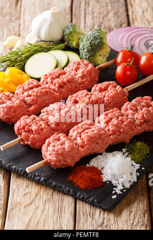 Raw, kofta kebab kofte da carni macinate su spiedini con ingredienti e verdure vicino sul piano verticale. Foto Stock