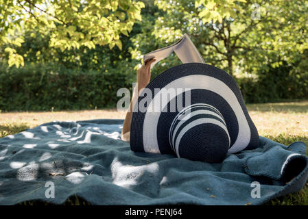 Donna sdraiata su un tappeto all'ombra di un albero di trascorrere una giornata di relax in estate la lettura di un libro. Foto Stock