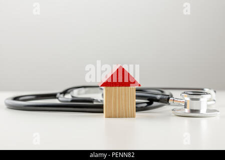 Immagine concettuale di un medico di uno stetoscopio giacente su una superficie bianca con un piccolo modello in legno di una casa con un luminoso tetto rosso con copyspace su grigio Foto Stock