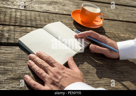 Primo piano delle mani di un uomo pronto a scrivere nella parte anteriore di un quaderno con pagine vuote accanto a una tazza di caffè. Foto Stock