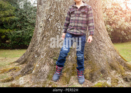 Elegante little boy in abiti casual jean e camicia si posa con grande albero in background. Foto Stock