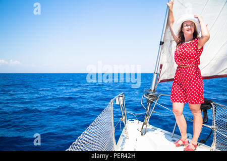 Femmina su barca a vela. Una giovane donna in un abito rosso e rosso scarpe e un cappello su uno yacht contro lo sfondo delle vele e il mare. Foto Stock