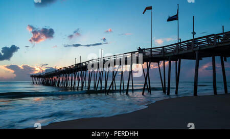 Un legno lungo ocean pier si estende sul mare verso un brillante sunrise come bandiere volare nella brezza. Foto Stock