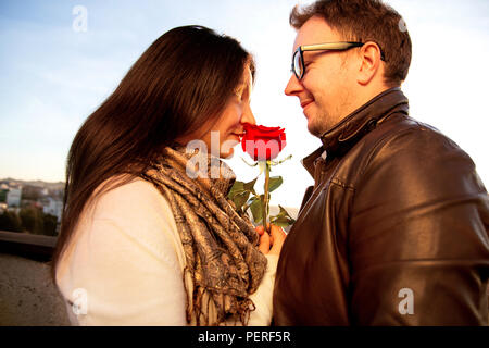 Coppia felice per il giorno di San Valentino. Amare giovane godendo il sole sul tetto e bella ragazza odori una rosa rossa, simbolo dell'amore. Foto Stock