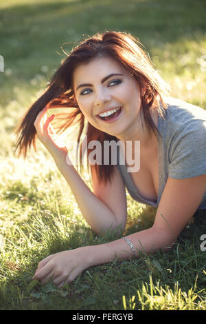 Closeup ritratto della bella sorridente giovane donna caucasica con rosso nero capelli, sdraiati sull'erba all'aperto, ridendo mostrando i denti, bellezza naturale la gioventù Foto Stock