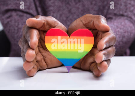 Close-up di una mano di un uomo proteggere striscia colorata cuore oltre la scrivania bianca Foto Stock