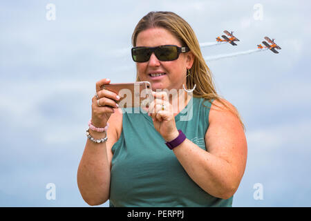 Donna prendendo selfie con iphone con wingwalkers nella distanza a Weymouth beach, Weymouth, Dorset Regno Unito su una calda giornata di sole nel mese di agosto Foto Stock