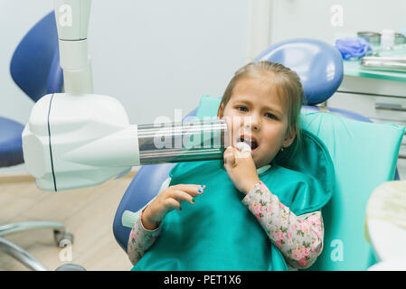 Bambino con una madre a un dentista di ricezione. La ragazza si trova in cattedra, dietro la madre. Il dottore lavora con un assistente. Procedura per la perforazione di un dente. Impostazione di una sigillatura temporanea. Foto Stock