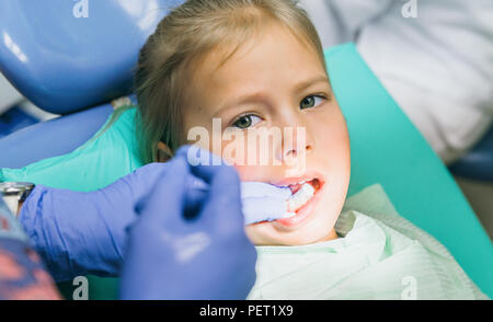 Bambino con una madre a un dentista di ricezione. La ragazza si trova in cattedra, dietro la madre. Il dottore lavora con un assistente. Procedura per la perforazione di un dente. Impostazione di una sigillatura temporanea. Foto Stock