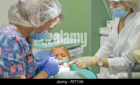 Bambino con una madre a un dentista di ricezione. La ragazza si trova in cattedra, dietro la madre. Il dottore lavora con un assistente. Procedura per la perforazione di un dente. Impostazione di una sigillatura temporanea. Foto Stock