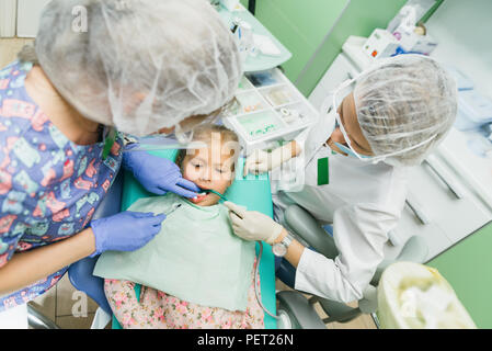 Bambino con una madre a un dentista di ricezione. La ragazza si trova in cattedra, dietro la madre. Il dottore lavora con un assistente. Procedura per la perforazione di un dente. Impostazione di una sigillatura temporanea. Foto Stock