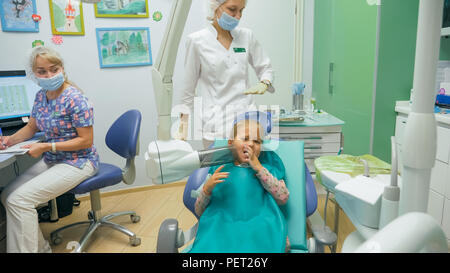 Bambino con una madre a un dentista di ricezione. La ragazza si trova in cattedra, dietro la madre. Il dottore lavora con un assistente. Procedura per la perforazione di un dente. Impostazione di una sigillatura temporanea. Foto Stock