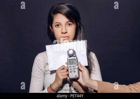 Ritratto di splendida latina ispanica bruna giovane donna in studio azienda white paper e misuratore di flash, luce di misurazione, su sfondo nero Foto Stock