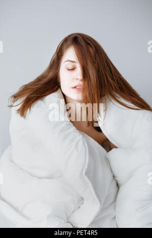 Ritratto di giovane e bella ragazza caucasica donna con capelli lunghi seduto nel letto avvolto coperto con coperta su sfondo bianco, sognando con chiuso Foto Stock