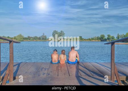 Bambini seduti sul molo. Tre bambini di differenti età - ragazzo adolescente, età elementare ragazzo e ragazza di età prescolare seduto su di un molo in legno. Estate e concetto di infanzia. I bambini sul banco a lago. I fratelli seduti sul molo in legno Foto Stock