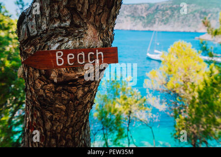 Assos village, Cefalonia. La Grecia. Spiaggia di legno segno di freccia su un pino che mostrano la direzione alla piccola spiaggia di nascosto. White boat in background sul mare Foto Stock