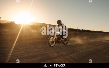 Biker maschio alla guida di una motocicletta vintage su strada sterrata. Uomo che cavalca veloce sulla sua bicicletta sulla strada di campagna. Foto Stock