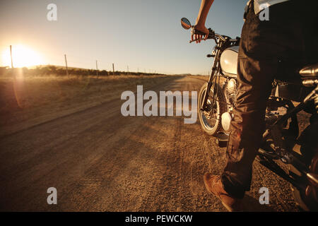 Colpo di un maschio di conducente in piedi sulla strada rurale con la sua moto. L uomo del paese ride con la sua motocicletta in piedi dalla strada. Bassa vista in sezione con copia sp Foto Stock