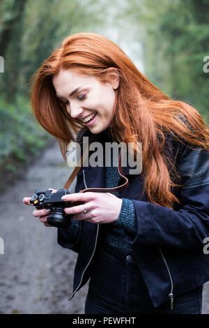Sorridente giovane donna con lunghi capelli rossi a piedi lungo una strada forestale, scattare foto con la fotocamera vintage. Foto Stock