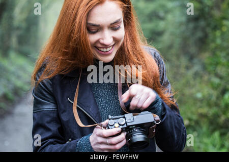 Sorridente giovane donna con lunghi capelli rossi a piedi lungo una strada forestale, scattare foto con la fotocamera vintage. Foto Stock