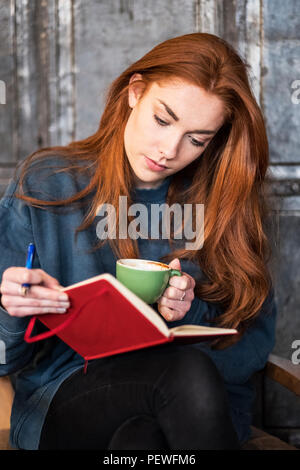 Giovane donna con lunghi capelli rossi seduta a tavola, tenendo notebook e la tazza di caffè. Foto Stock