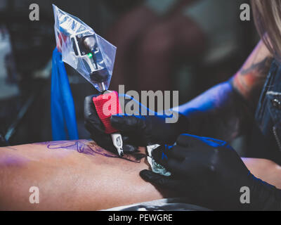 Vista ravvicinata del tatuaggio artista dimostra il processo di getting black skull tattoo con vernice. Femmina concentrato la colorazione di inchiostro di contorno sulla gamba con la pistola. Luce al neon. Foto Stock