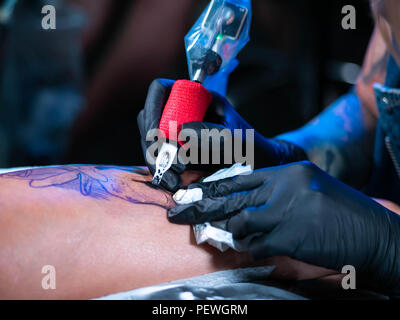 Vista ravvicinata del tatuaggio artista dimostra il processo di getting black skull tattoo con vernice. Femmina concentrato la colorazione di inchiostro di contorno sulla gamba con la pistola. Luce al neon. Foto Stock