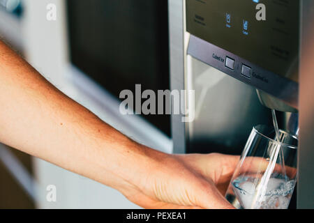 Mano maschio è versando acqua fredda e cubetti di ghiaccio dal distributore del frigorifero domestico. Foto Stock