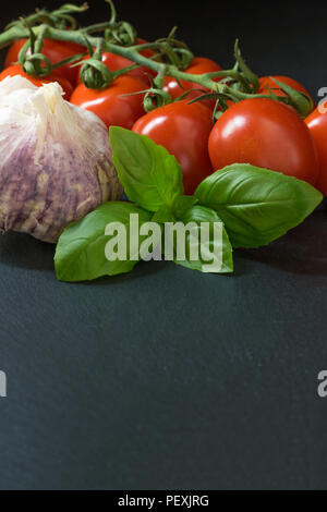 Tasto basso colpo di arbusti di pomodoro, basilico e aglio sono giacenti sulla pietra nera table.Tutto su sfondo scuro. Verticalmente. Foto Stock