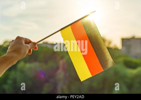 Mano trattiene bandiera della Germania una finestra aperta. Lo sfondo è blu cielo, la silhouette della città, il tramonto. Foto Stock