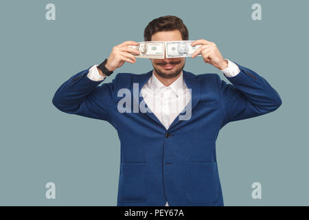 Bello happy businessman in giacca blu e camicia bianca permanente e la holding e coperto molti dollari davanti agli occhi e sorridente . Indoor, studio Foto Stock