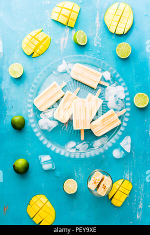Mango popsicles su una lastra di vetro con cubetti di ghiaccio, lime e mango fruit,vista superiore, scheda blu. Foto Stock