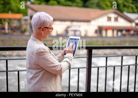 Vista posteriore foto di senior donna in piedi sul ponte e cercando il modo in Navigator. donna legge il libro guida. femmina è guardando la mappa su Foto Stock