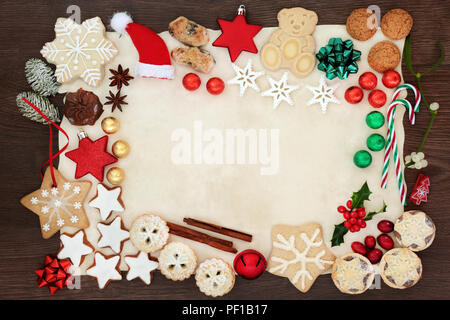 Festa di Natale del bordo dello sfondo compresi addobbi per l'albero, biscotti, dolci, cioccolatini, spezie e flora invernale su carta pergamena sul rovere rustico. Foto Stock