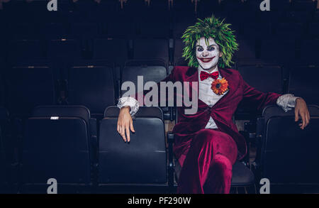 Un maschio cosplayer vestito come il burlone di Batman e DC Comics franchising seduto da solo in un abbandonato movie theater e guardando minaccioso Foto Stock