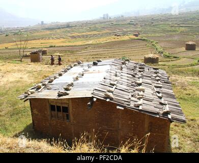 Due donne in tradizionali panni bhutanesi camminare nei pressi di una vecchia casa di argilla o terra rifugio Casa con pietre sul tetto nel piccolo villaggio a Punakha Foto Stock