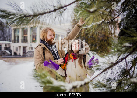 Giovani amare giovane uomo caucasico biondo con i capelli lunghi e la barba, donna con sorriso toothy divertirsi, indulgere, imbrogliamo intorno winter park vicino a Natale tr Foto Stock