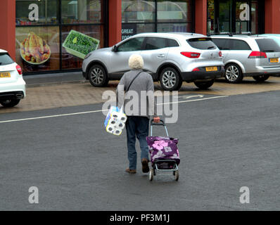 Vecchia donna con il carrello che porta rotoli di carta igienica del parco auto verso il pesce e patatine fritte piselli annunci grande povertà area Drumchapel, Glasgow, Regno Unito Foto Stock