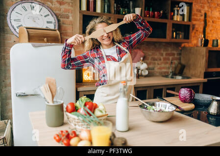 Giocoso casalinga in un grembiule copre gli occhi con le spatole in legno, cucina interno su sfondo. Felice cuoca facendo una sana alimentazione vegetariana, Foto Stock