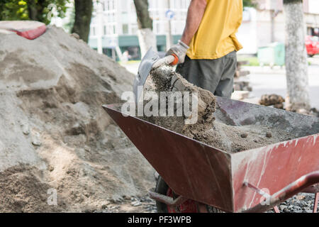 Lavoratore usare la pala e riempire la carriola con sabbia per la costruzione della strada sul marciapiede. Strumenti di costruzione. La pala e il carrello su ruote con sabbia sulla costruzione di strade Foto Stock