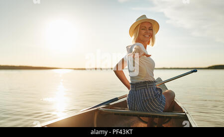 Sorridente giovane donna guardando indietro sopra la sua spalla e raggiungere con la mano mentre paddling una canoa sulle sponde di un lago in estate Foto Stock
