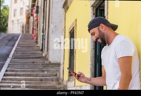 Ritratto di un uomo spagnolo con la barba guardando sul suo cellulare, sorridente, indossando un cappello snapback e una t-shirt, all'esterno. Foto Stock