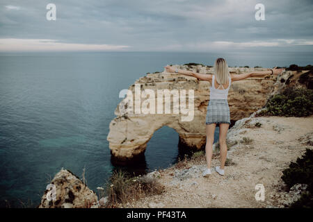 Giovane donna turista gode di splendide vedute dell'Oceano Atlantico e il paesaggio al largo in Portogallo e alza le braccia verso l'alto mostra ho Foto Stock