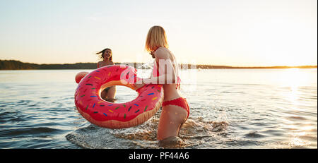 Sorridente ragazza in un bikini in possesso di un anello di nuotare e spruzzi il suo amico con acqua mentre godendo un pomeriggio in un lago Foto Stock