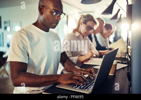 Giovani designer africani incentrata sul suo lavoro mentre è seduto in una fila con i colleghi a una tabella in un ufficio moderno Foto Stock